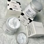 کرم شیر گاو ایباگ EBUG با بسته‌ بندی سفید و طرح پوست گاو، حاوی کرم روشن‌کننده و آبرسان برای پوست صورت و بدن، نمایش با درب باز و بافت نرم و کرمی محصول